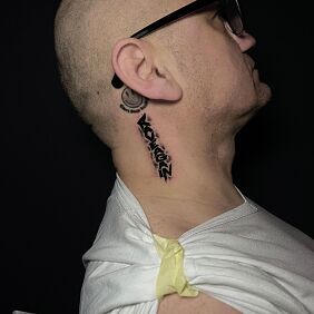 Tatuaż Napis "RAVEAGAIN" za uchem motyw: czarno-szare styl: graficzne / ilustracyjne